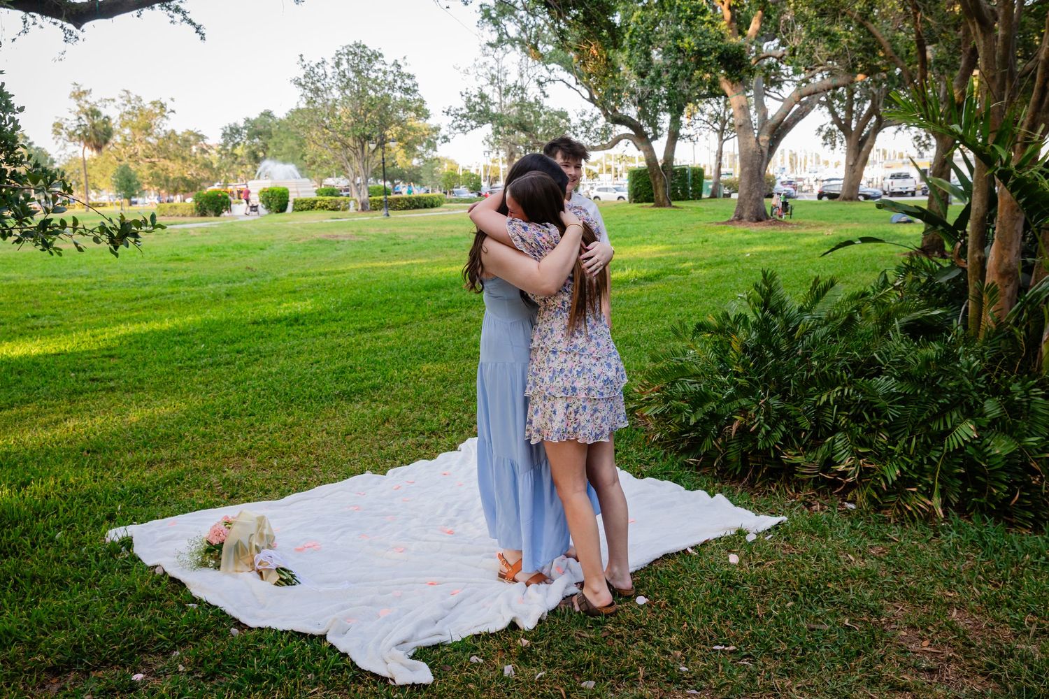 Two women share a heartfelt hug outdoors on a white blanket in a lush green park setting.