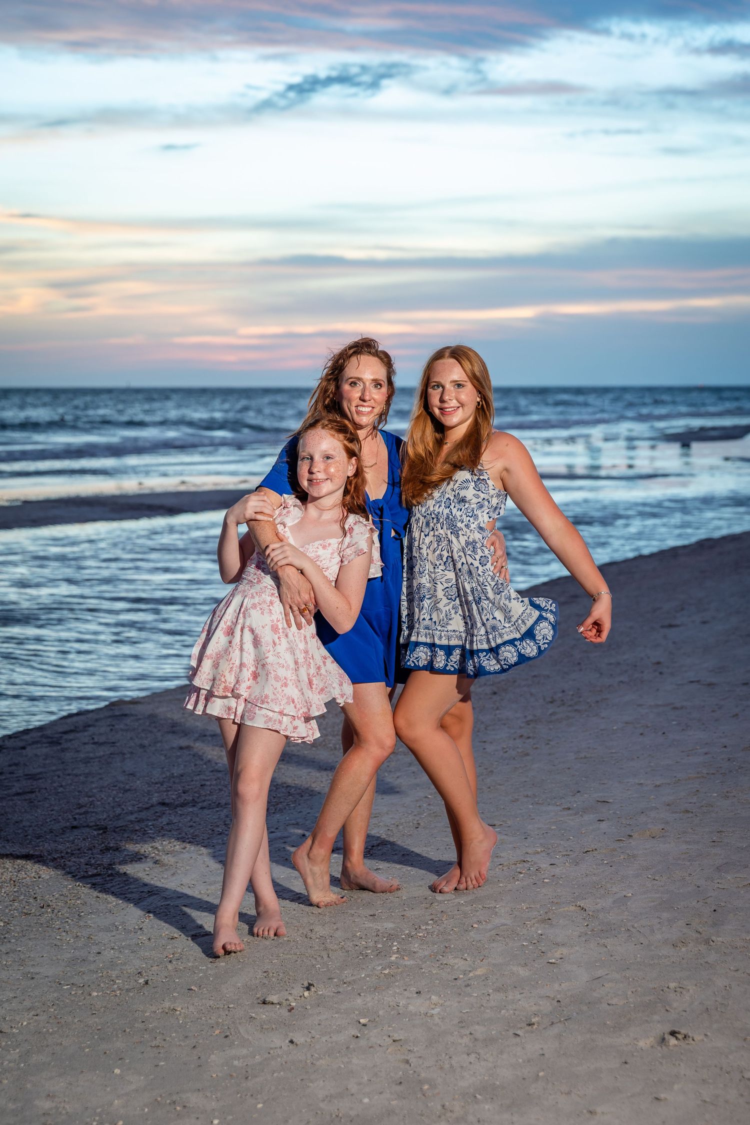 Three females pose barefoot on a sandy beach at sunset, wearing colorful summer dresses with ocean waves behind them.