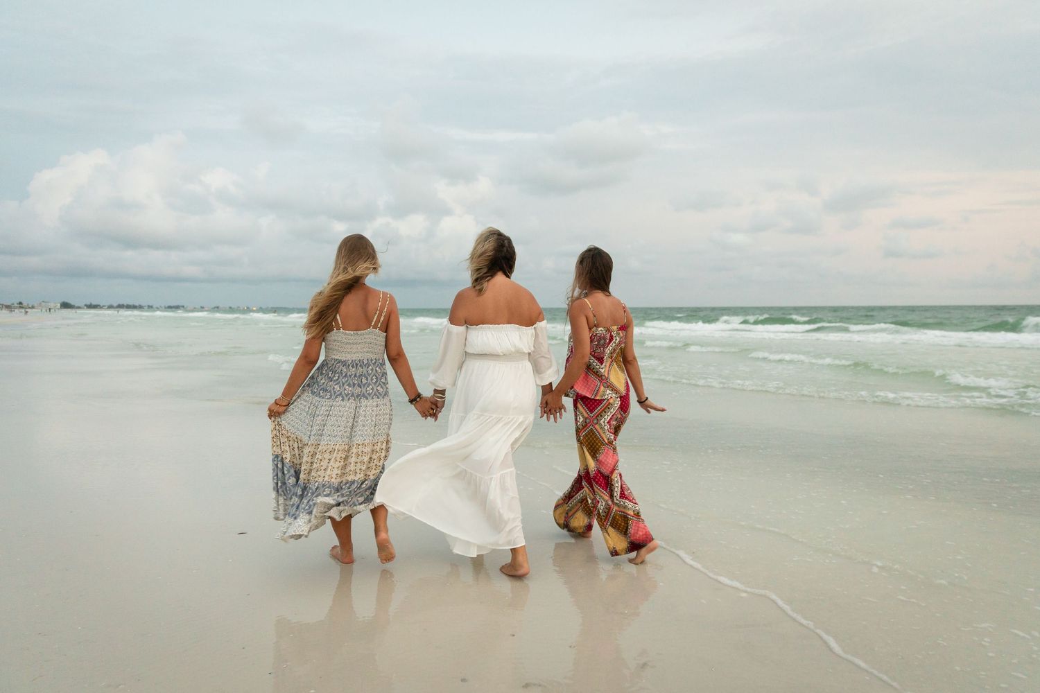 Three women in flowing dresses walk hand-in-hand along a serene beach with gentle waves and cloudy skies.