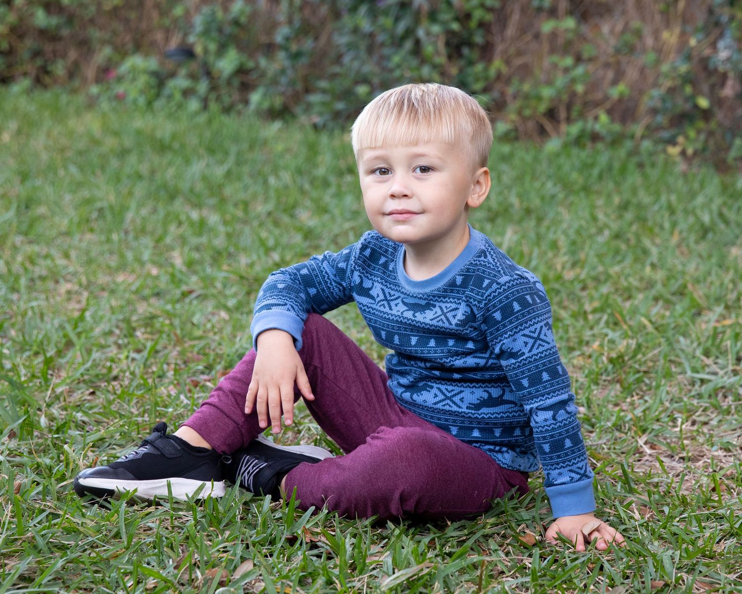 A young blonde toddler boy sits cross-legged on green grass wearing a blue patterned sweater and burgundy pants.