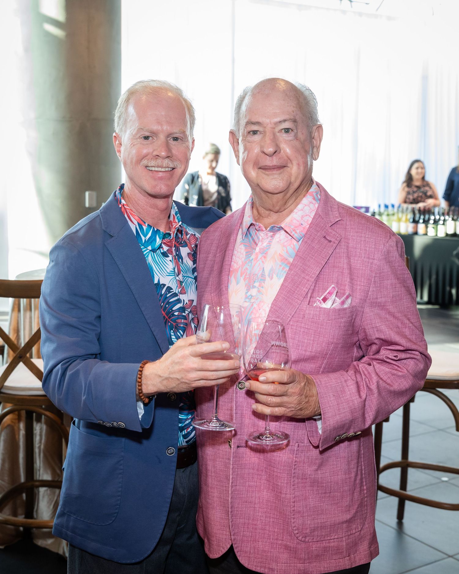 Two men in colorful blazers smile and clink wine glasses at an elegant indoor social event.