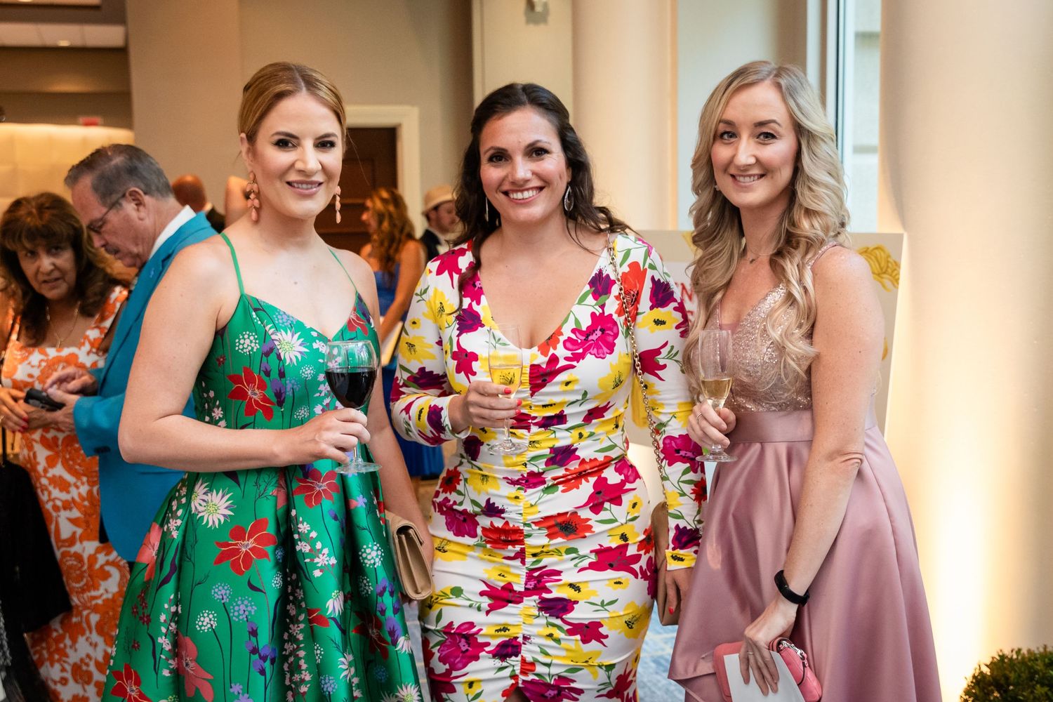 Three women in floral dresses smiling and holding drinks at an elegant indoor social event.