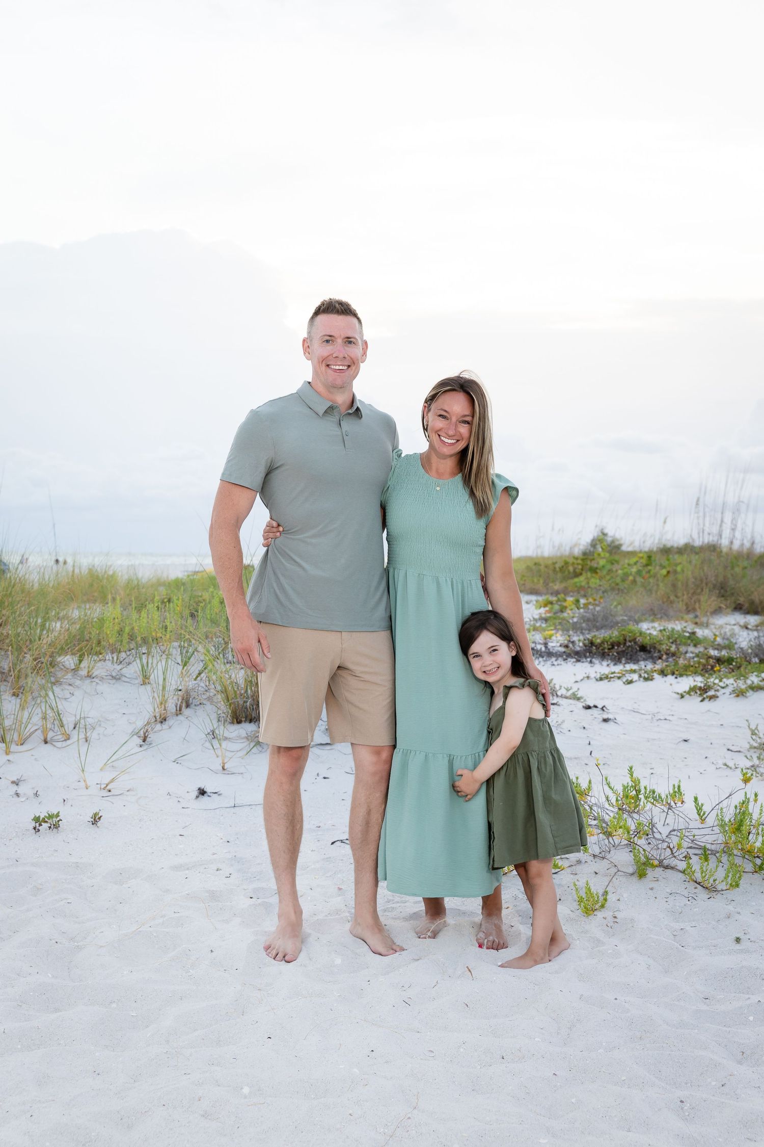 Family of three posing on white sand beach, parents in green outfits with young daughter, coastal dunes background.