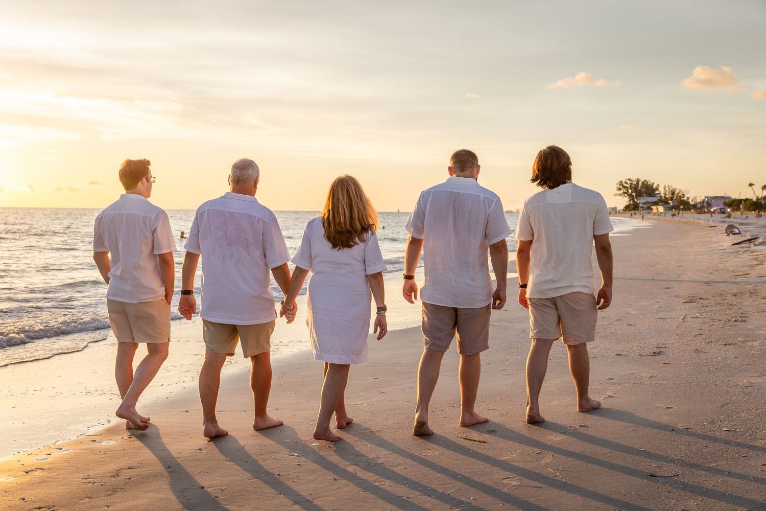Five people in white shirts walk along a sunny beach at sunset, viewed from behind.