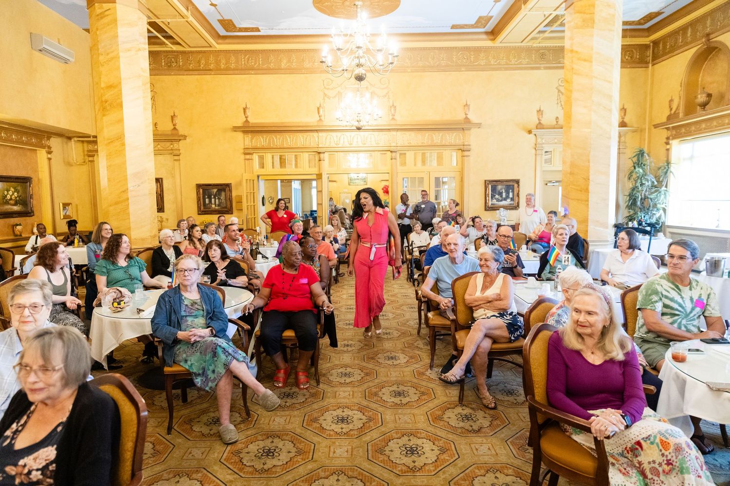A model in a pink dress walks the runway at an elegant indoor fashion show with seated audience.