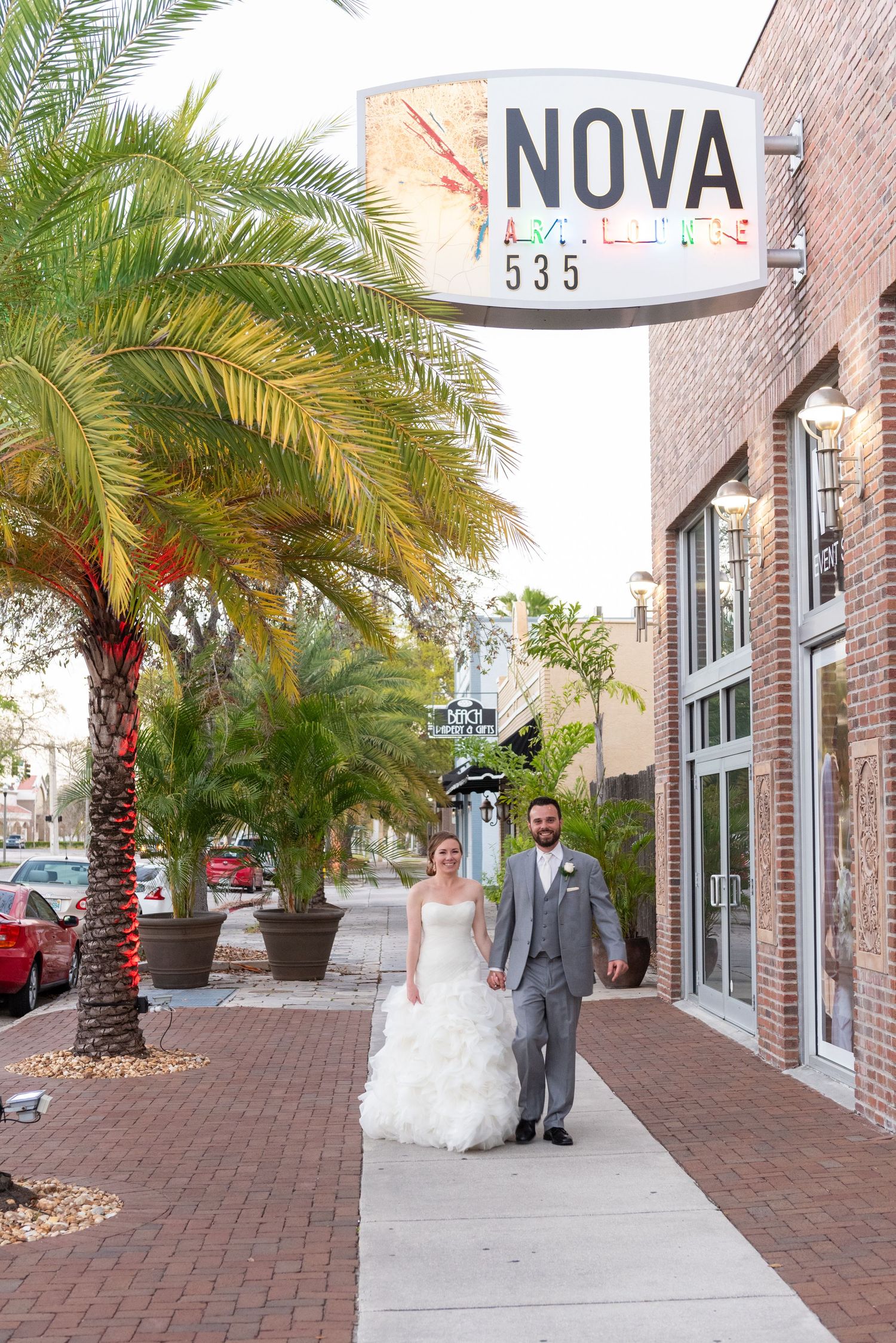Bride and groom walk hand in hand on sidewalk beneath Nova Art Lounge sign at 535 during wedding.