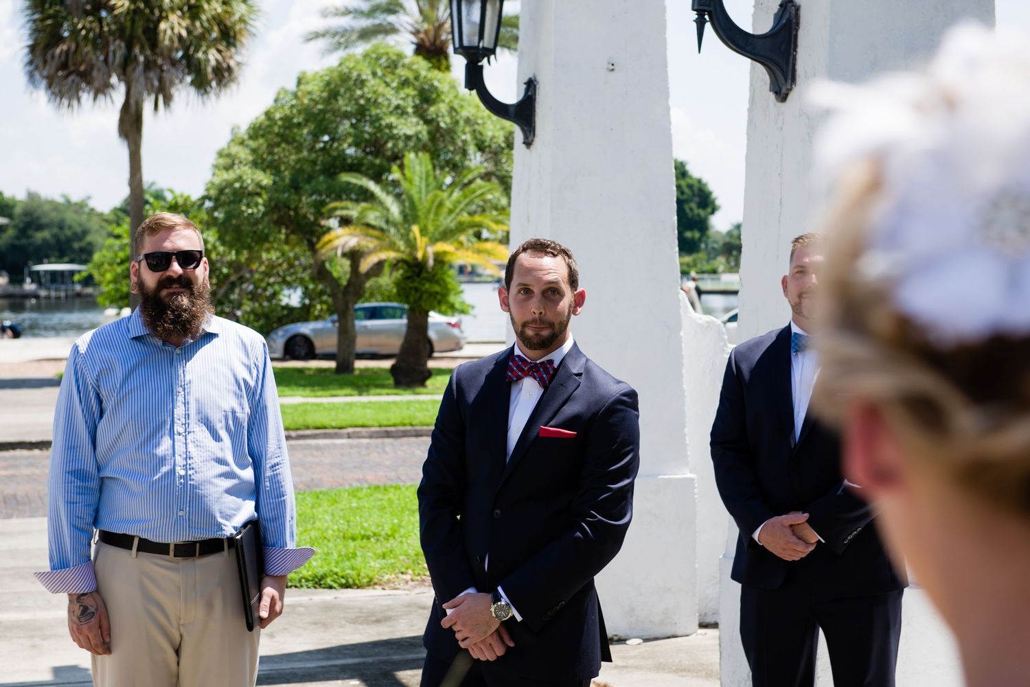 Groom in navy tuxedo with bow tie stands with groomsmen outdoors near palm trees at a sunny wedding ceremony.