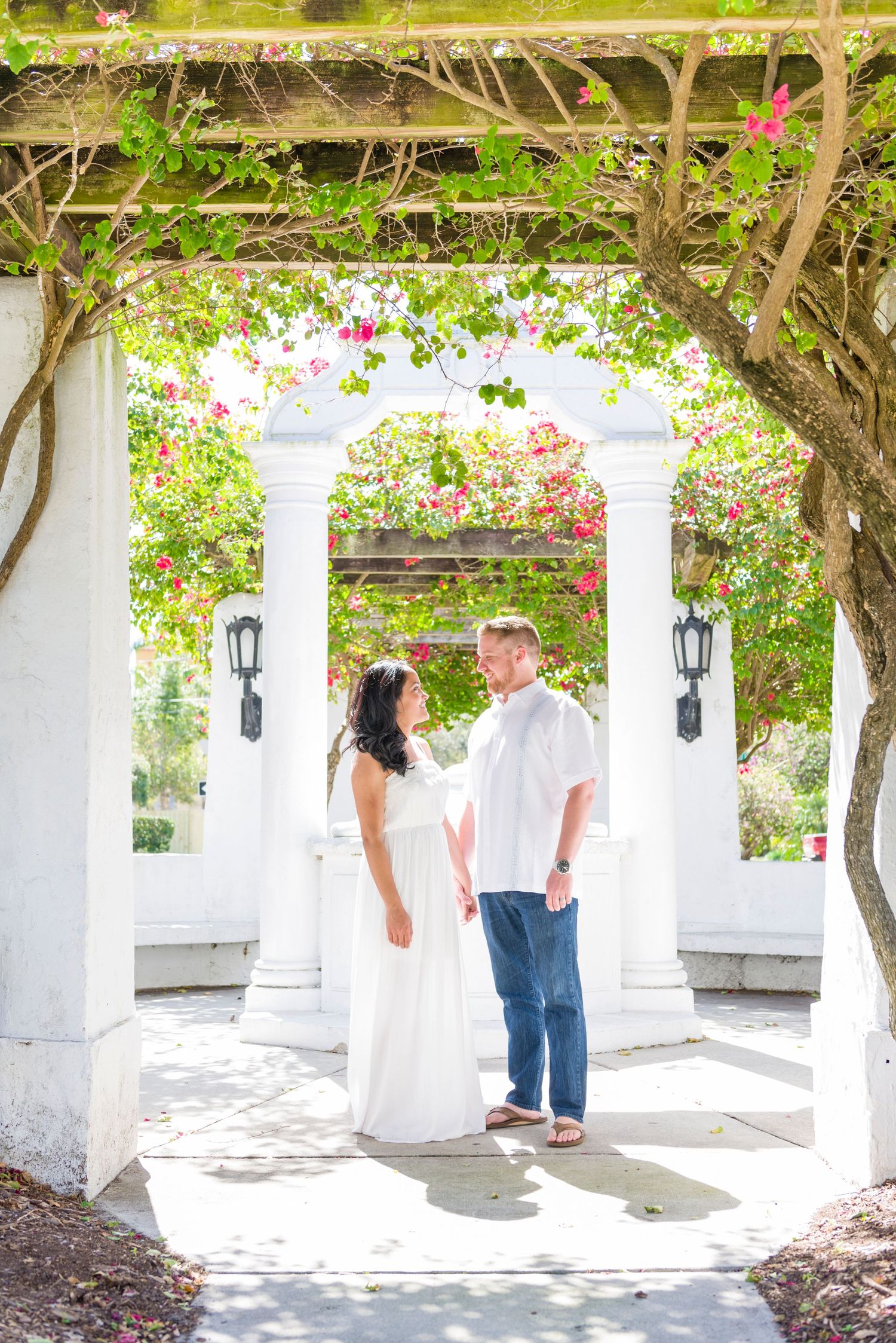 Couple in white dress and jeans standing under floral pergola with white columns and pink bougainvillea blooms.