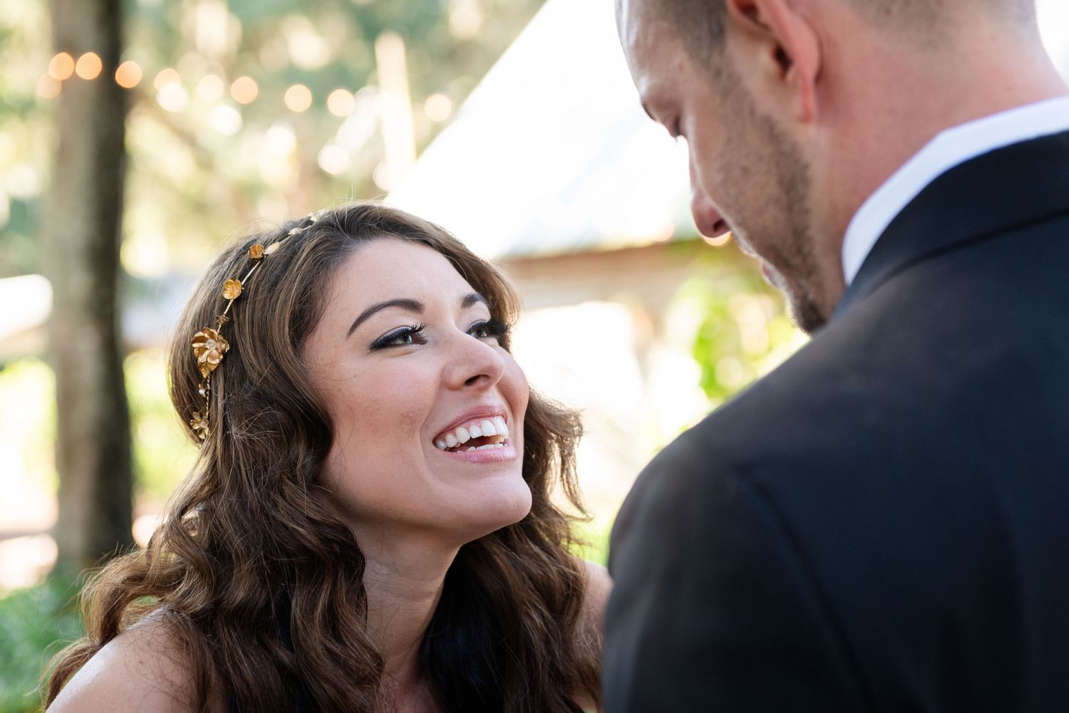 A radiant bride smiles joyfully at her groom during an outdoor wedding ceremony, wearing a floral hair accessory.