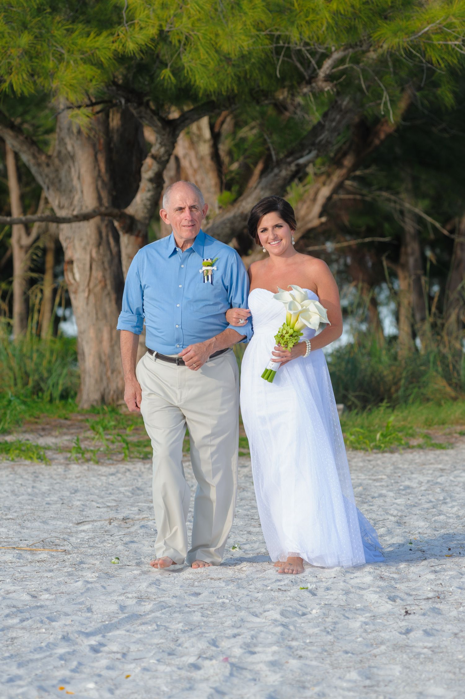 Bride in white gown holding bouquet walks with older man in blue shirt on sandy beach with trees.