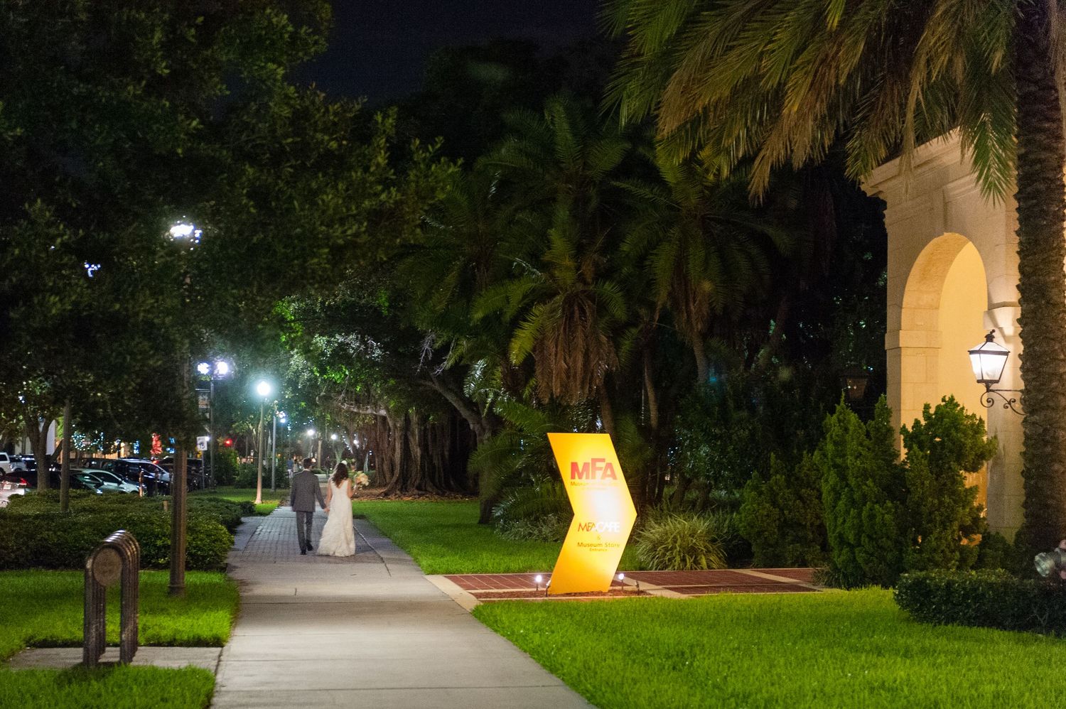 Bride and groom stroll along palm-lined pathway outside venue at night.