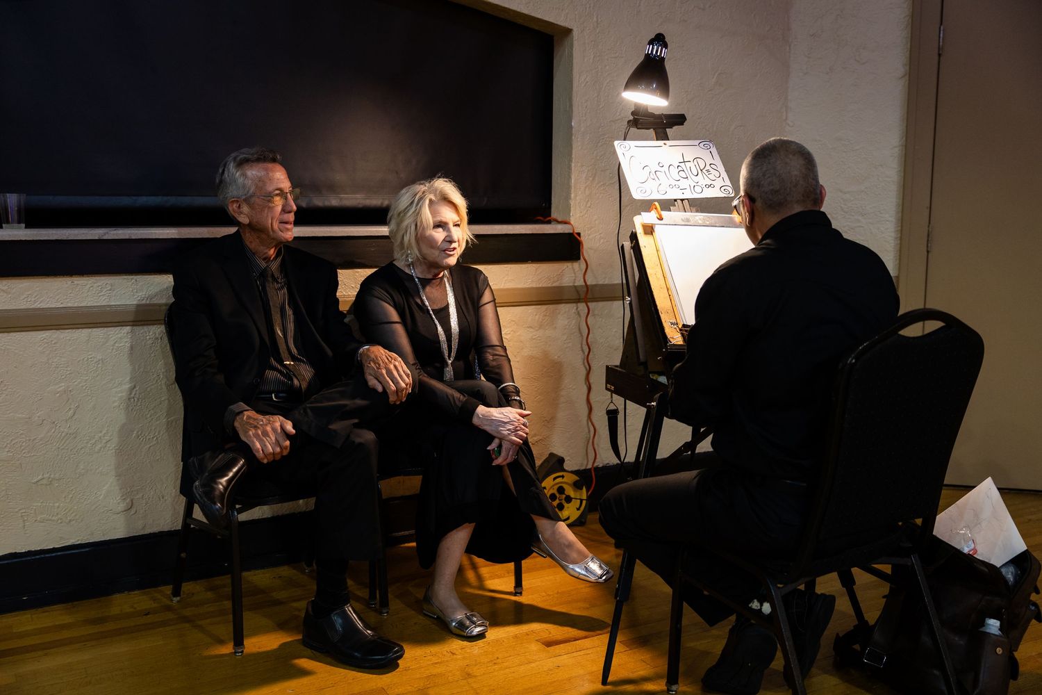 Two people sit and converse with an artist sketching at an easel in a dimly lit studio setting.