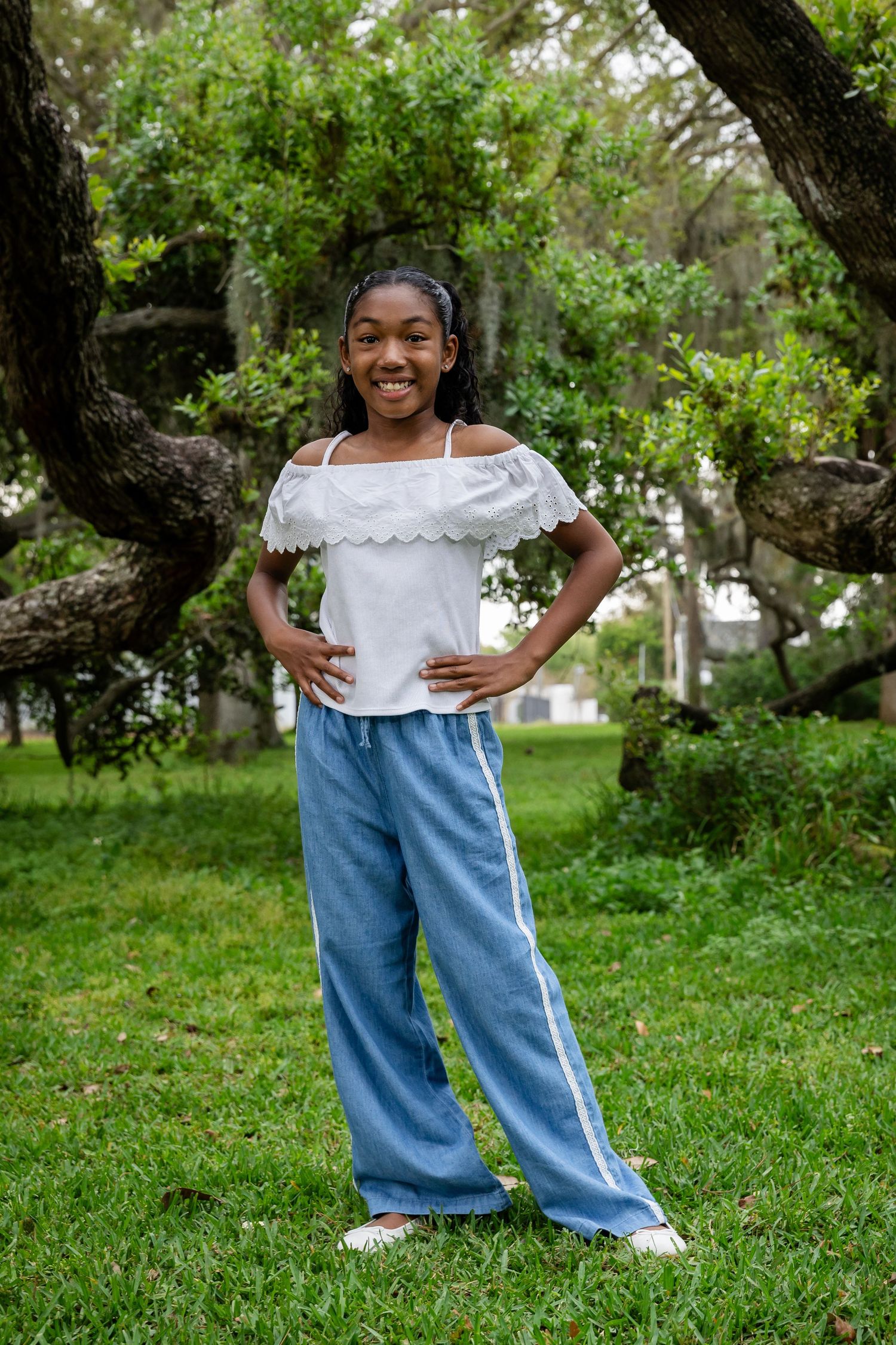 Smiling young Black girl posing with hands on hips wearing white off-shoulder top and blue wide-leg pants in a park.