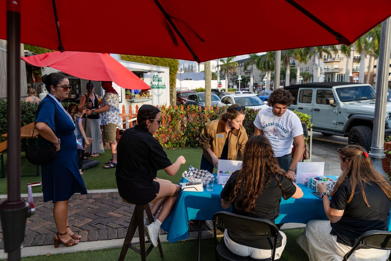 People gathered at outdoor tables under red umbrellas at a street event in a sunny urban setting.