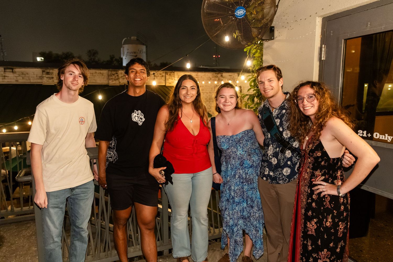 Group of six young adults smiling together on a rooftop bar patio at night with string lights.