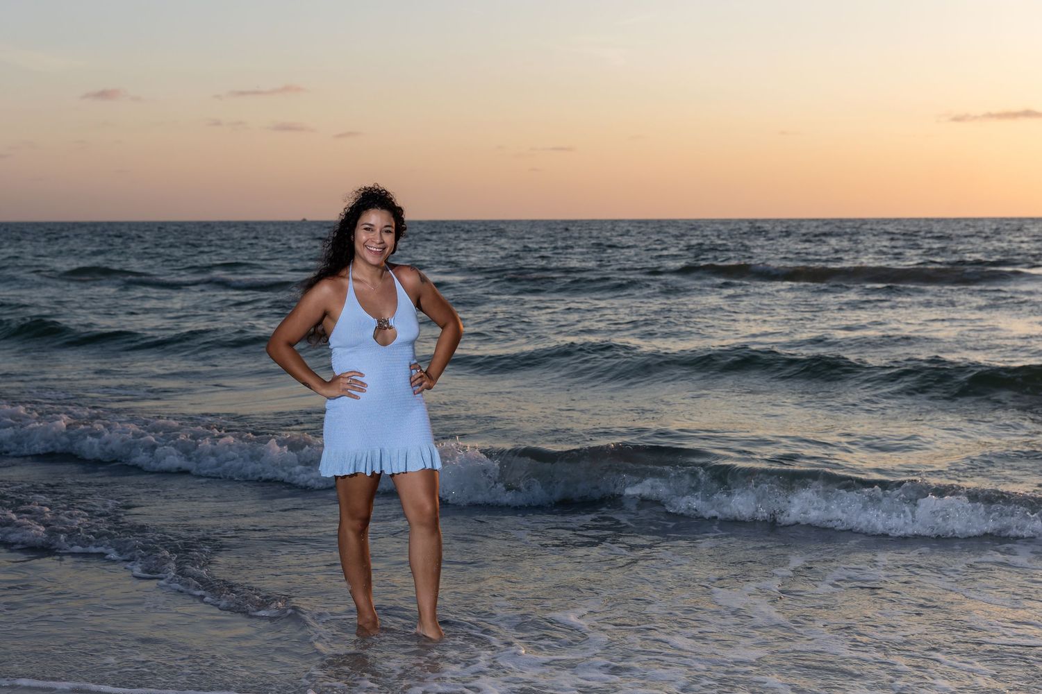 Woman in white dress posing on beach at sunset with ocean waves behind her.