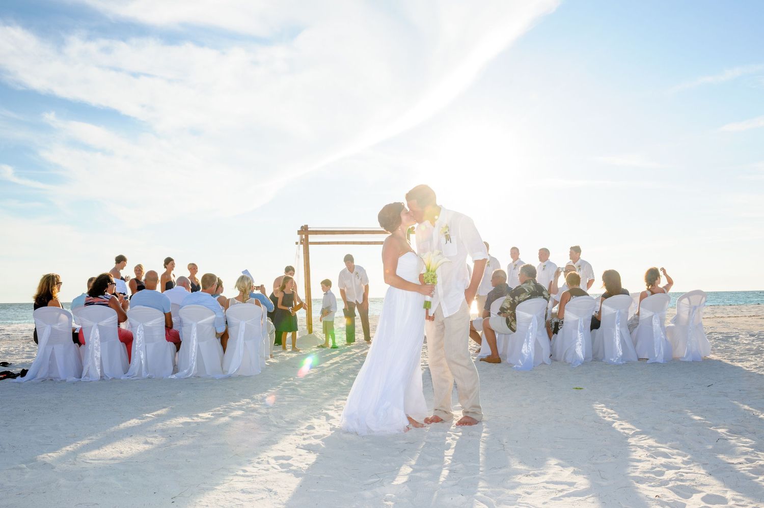 Newlyweds share first kiss on white sand beach with wedding party and guests behind them.