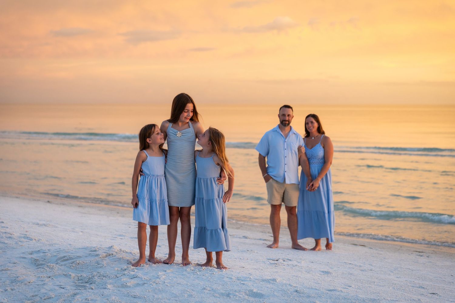 Family of five in matching blue outfits poses on a beach at golden sunset with calm ocean waves behind them.