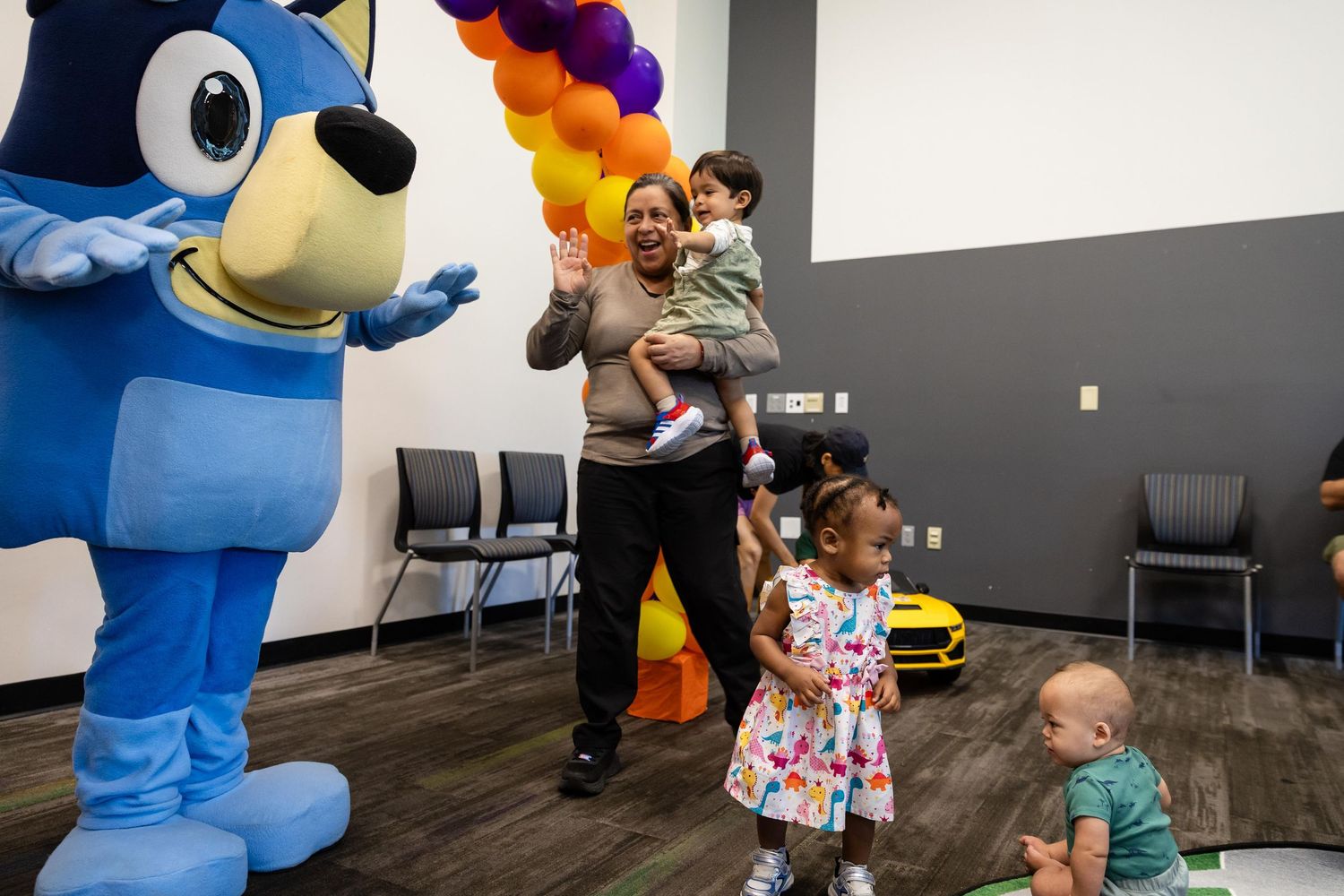 Family celebrates with Bluey mascot at colorful balloon arch party with children playing nearby.