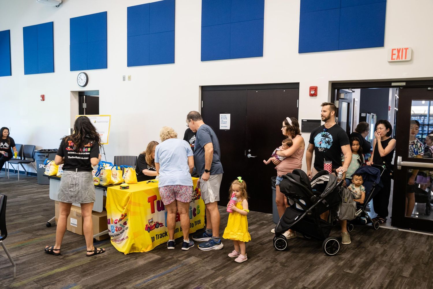 Families gather at an indoor community event with a yellow information table in a modern recreation center.