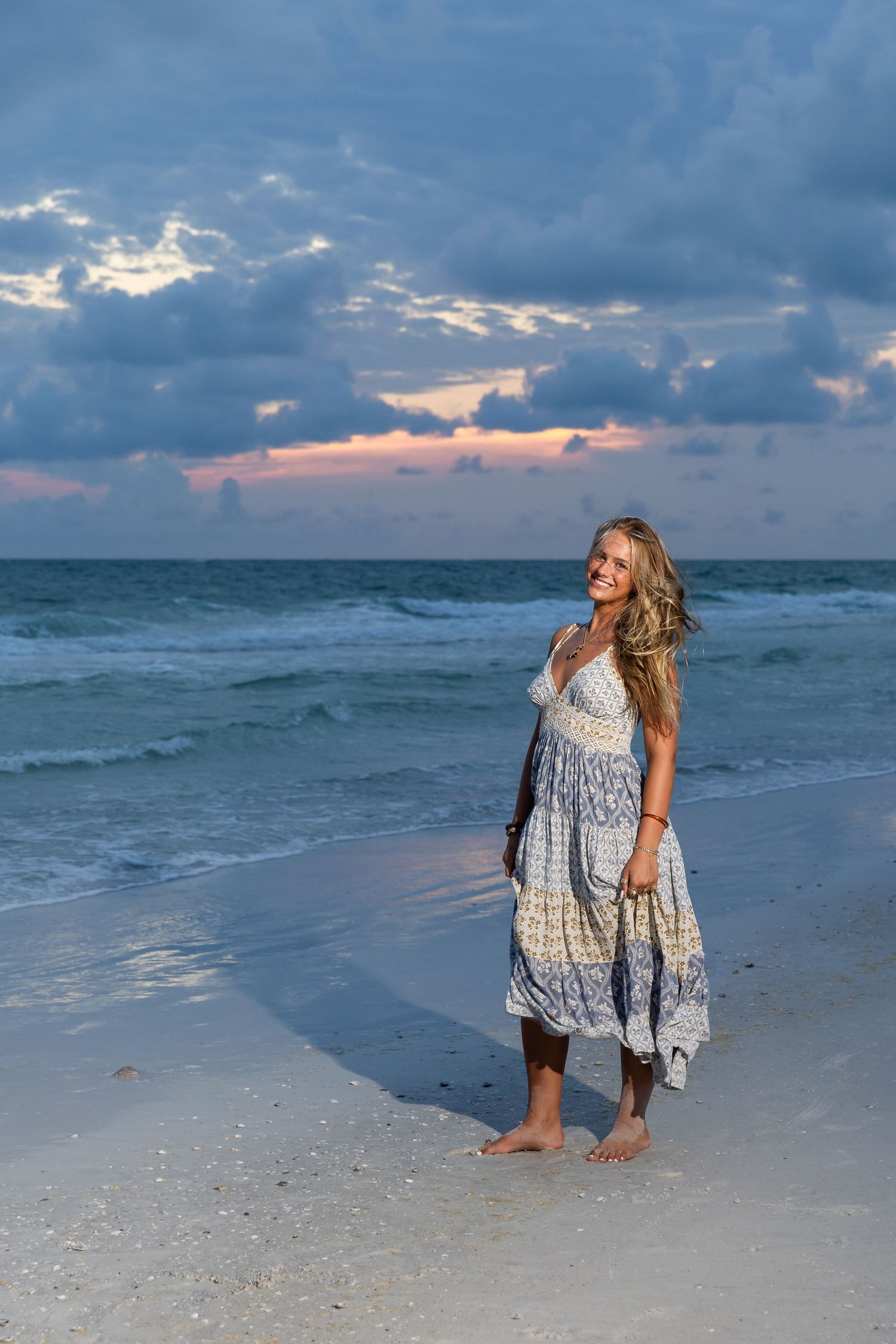 Woman in boho dress standing barefoot on sandy beach at sunset with dramatic cloudy sky and ocean waves.