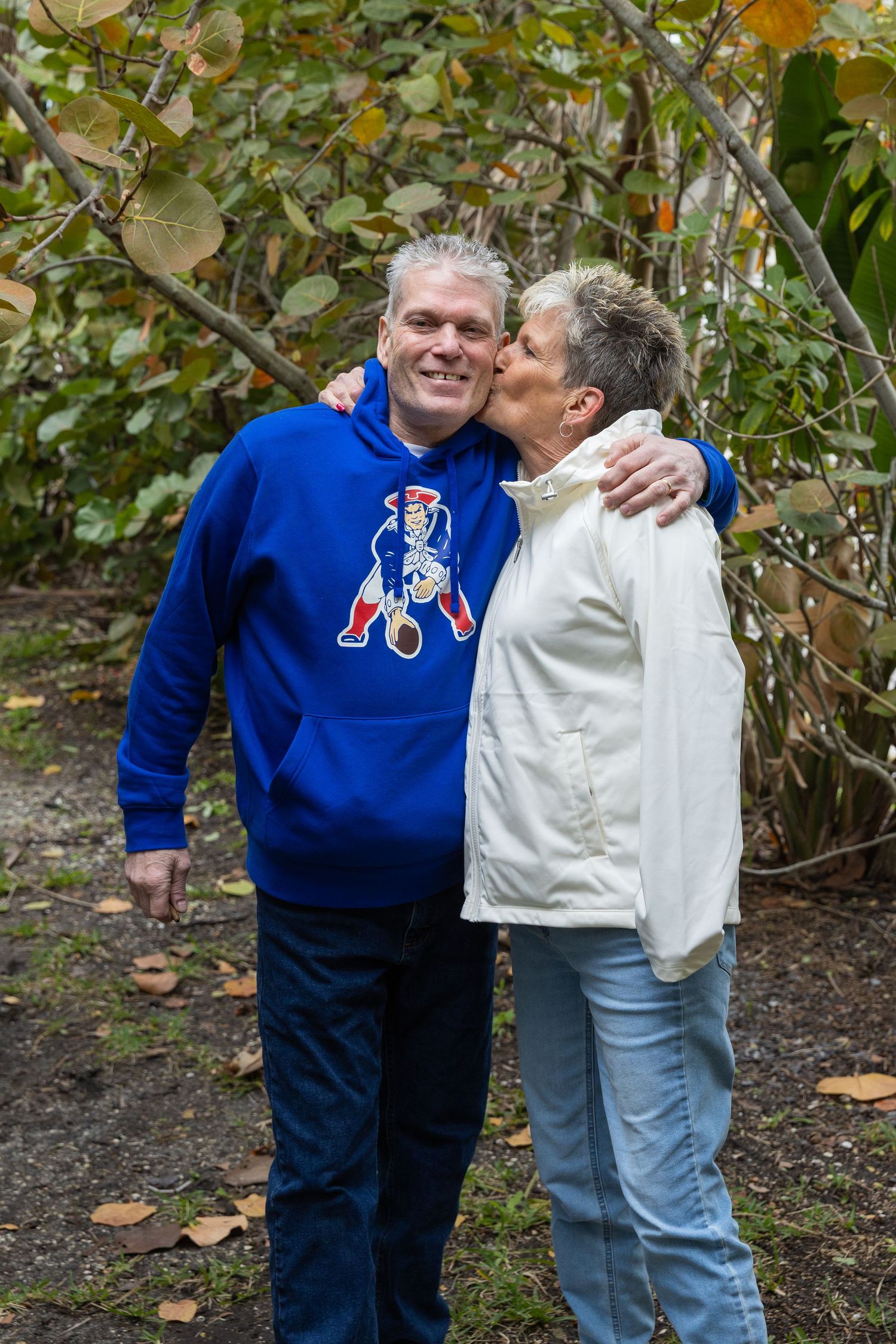 A smiling man in a New England Patriots hoodie receives a kiss on the cheek from a woman outdoors.