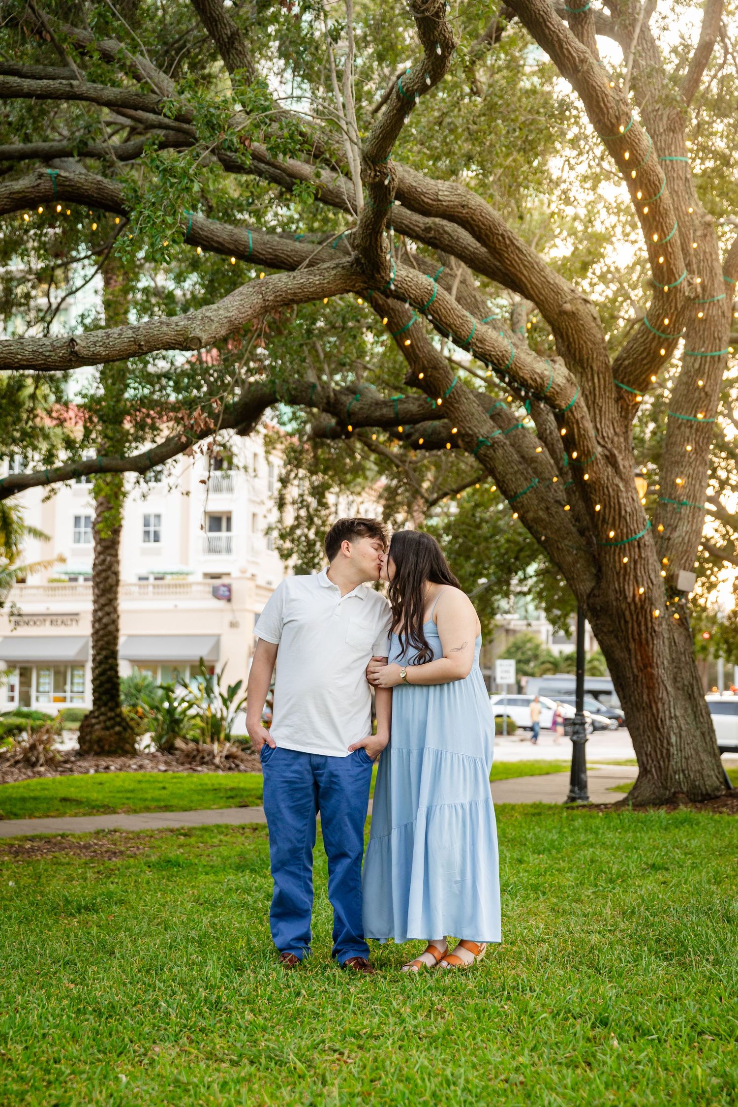 A couple shares a tender kiss under a large oak tree with string lights in a lush green park at golden hour.