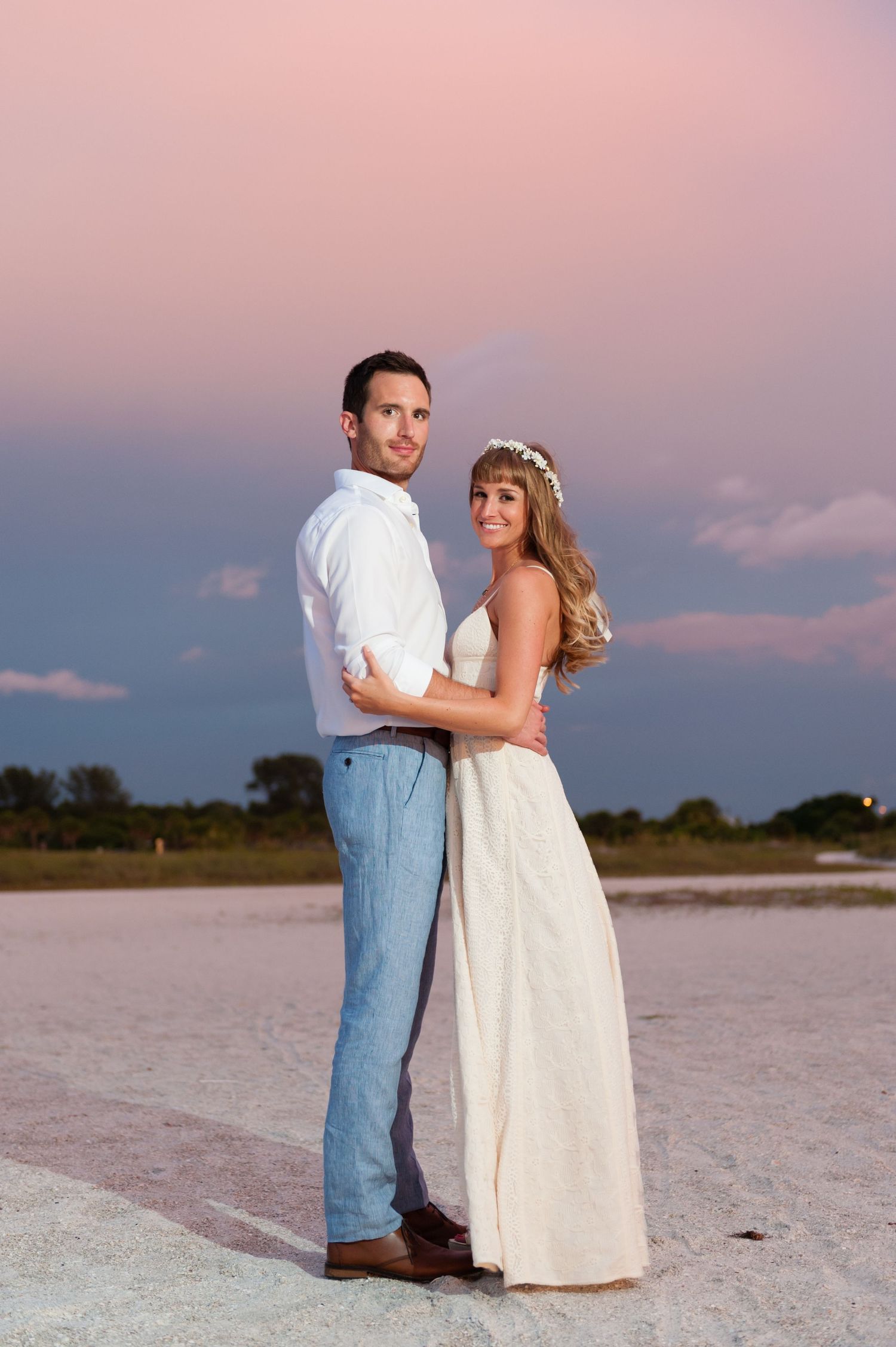 Couple in wedding attire embracing at sunset on a salt flat, woman in white gown with floral crown, man in white shirt.