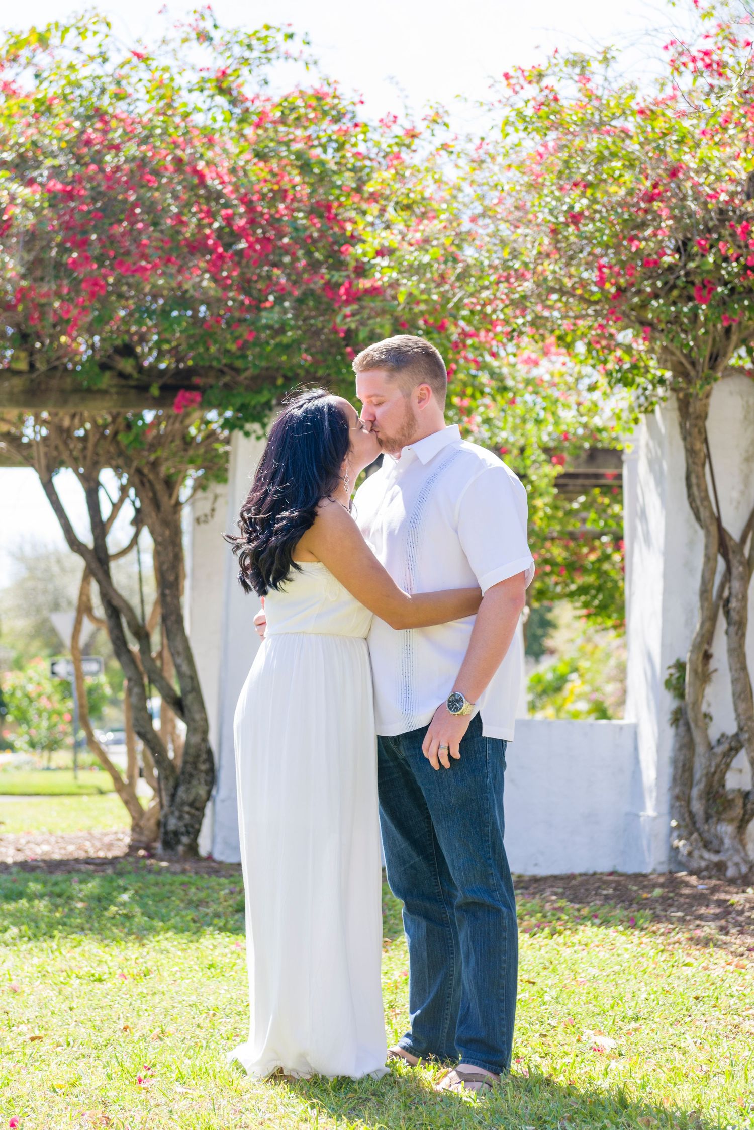 Couple sharing a romantic kiss outdoors under a flowering bougainvillea arbor, woman in white dress, man in white shirt.