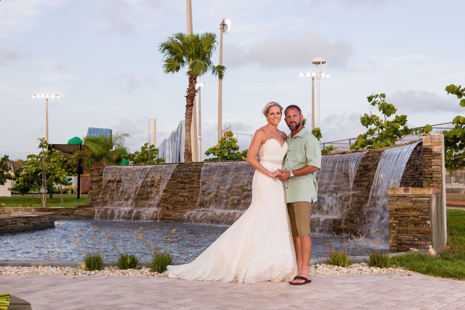 Bride and groom posing in front of a waterfall feature at an outdoor wedding venue with palm trees.