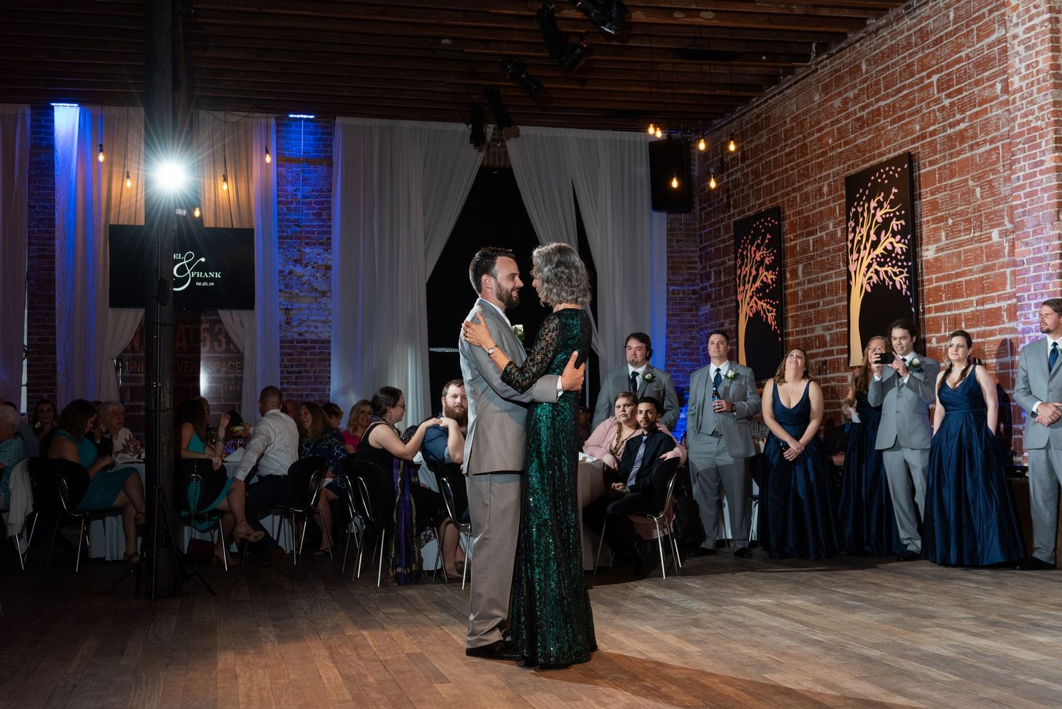 Groom shares emotional mother-son dance in elegant brick-walled wedding reception venue with guests watching.
