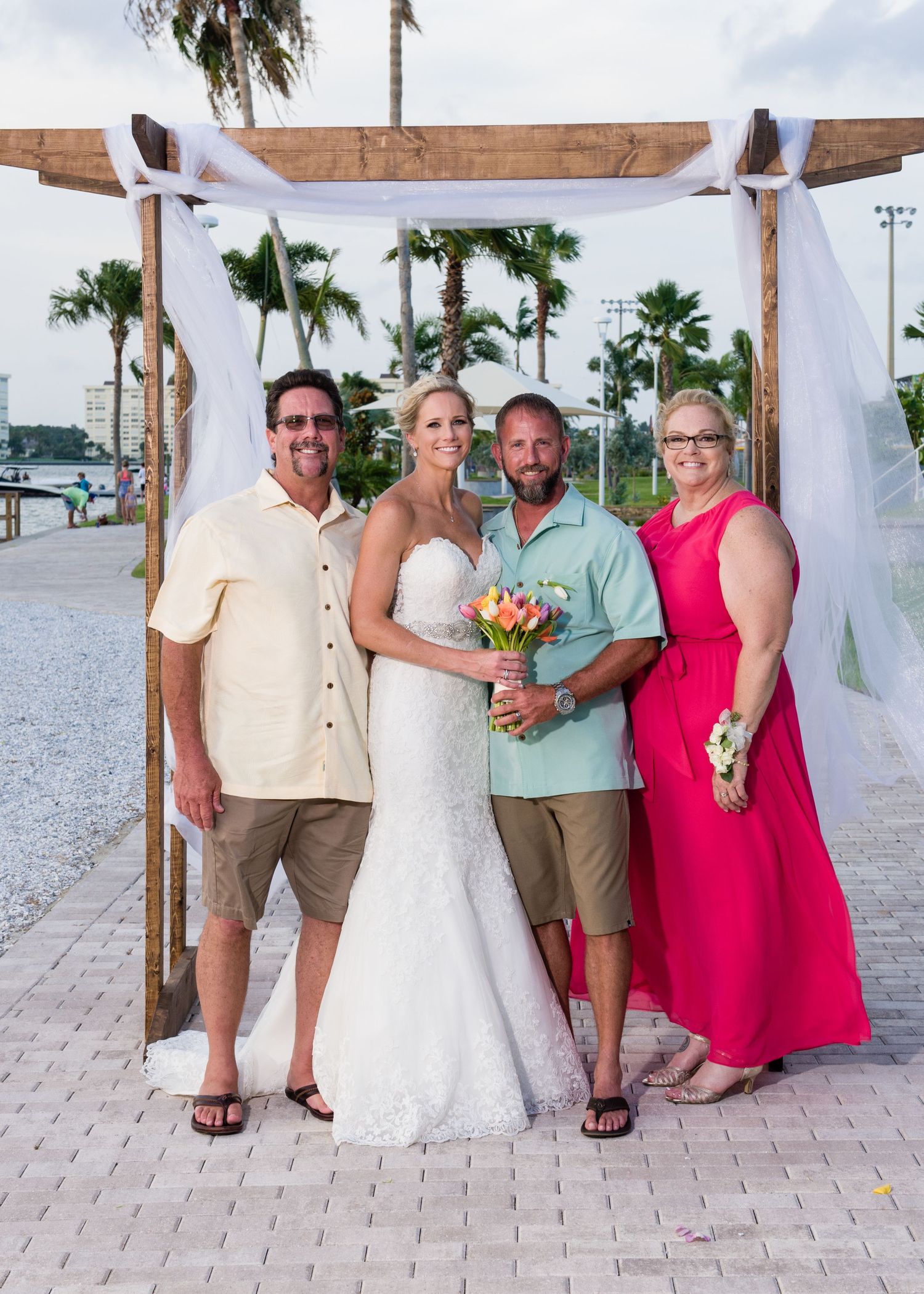 A bride in a white gown poses with three guests under a wooden arch at an outdoor waterfront wedding.