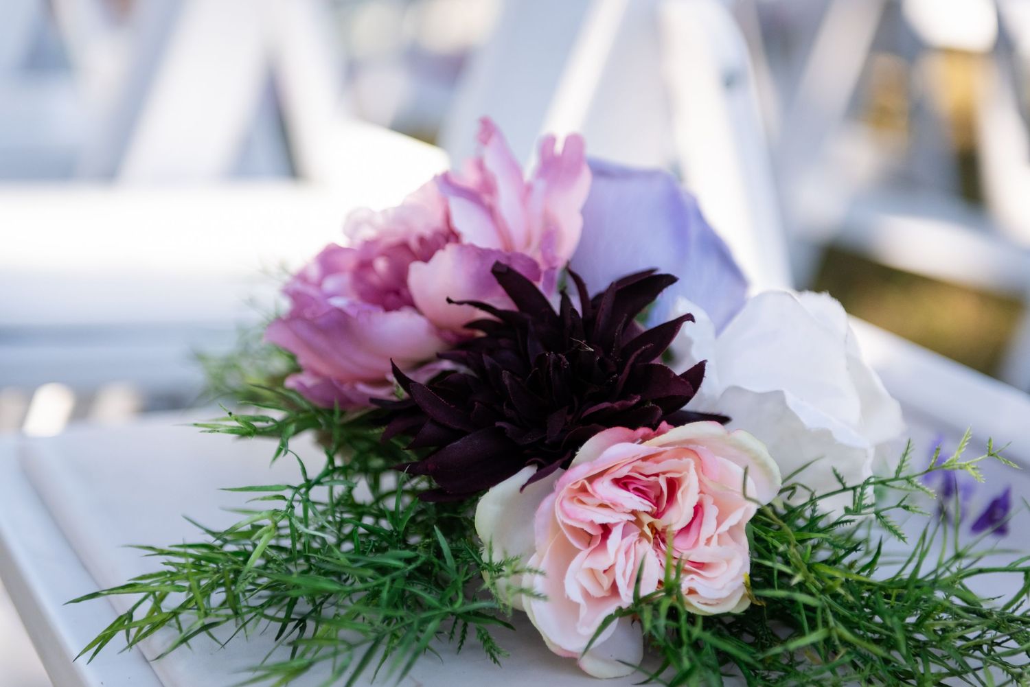 Pink peony, dark dahlia, and blush carnation floral arrangement with greenery on white chair at outdoor wedding.