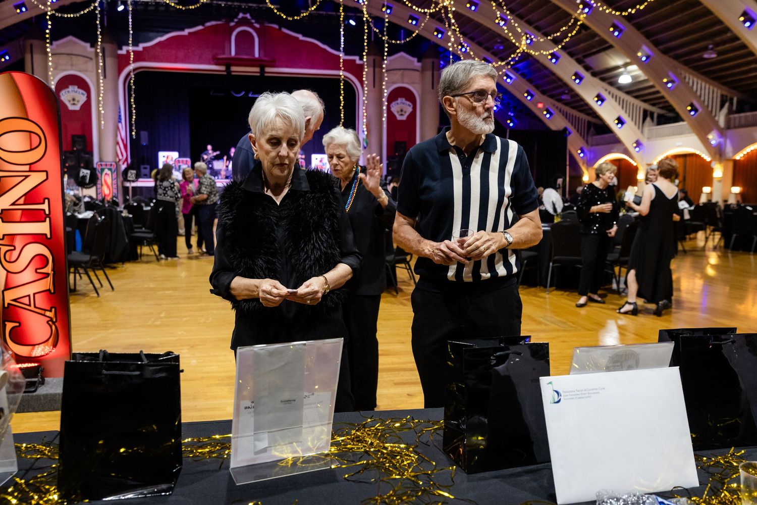 Two guests examine items at a casino-themed charity gala event in a decorated ballroom.