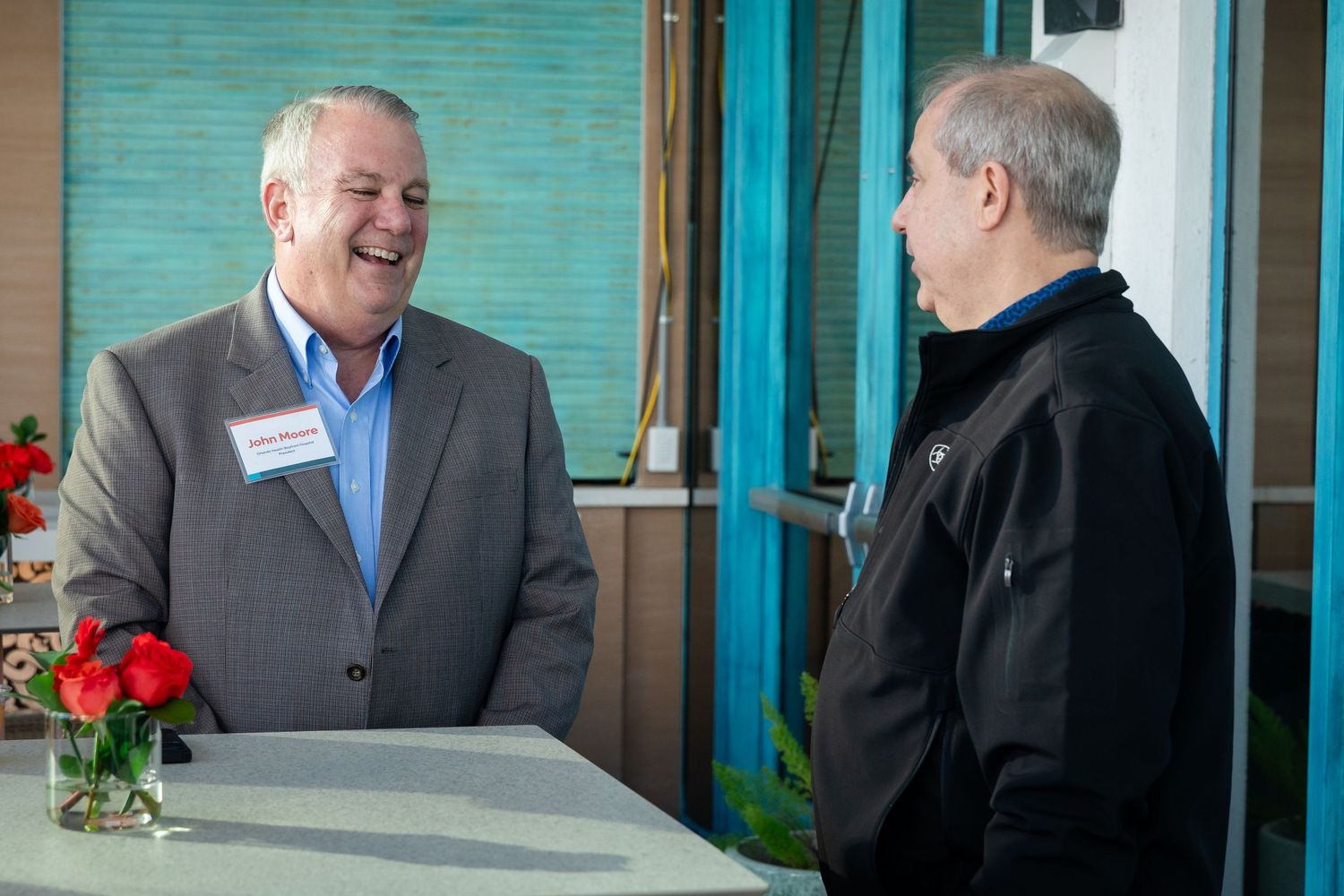 Two men in business casual attire smile and converse at a high-top table during a professional networking event.
