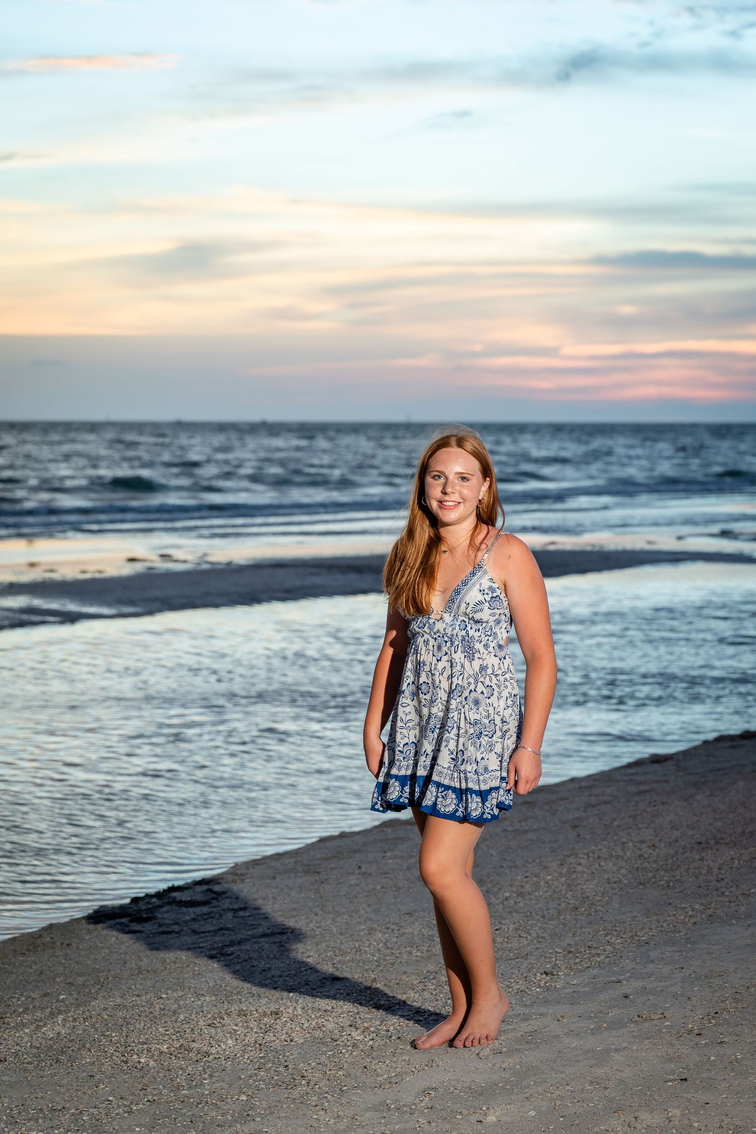 Young blonde woman in blue floral sundress standing barefoot on sandy beach at sunset with ocean waves behind her.