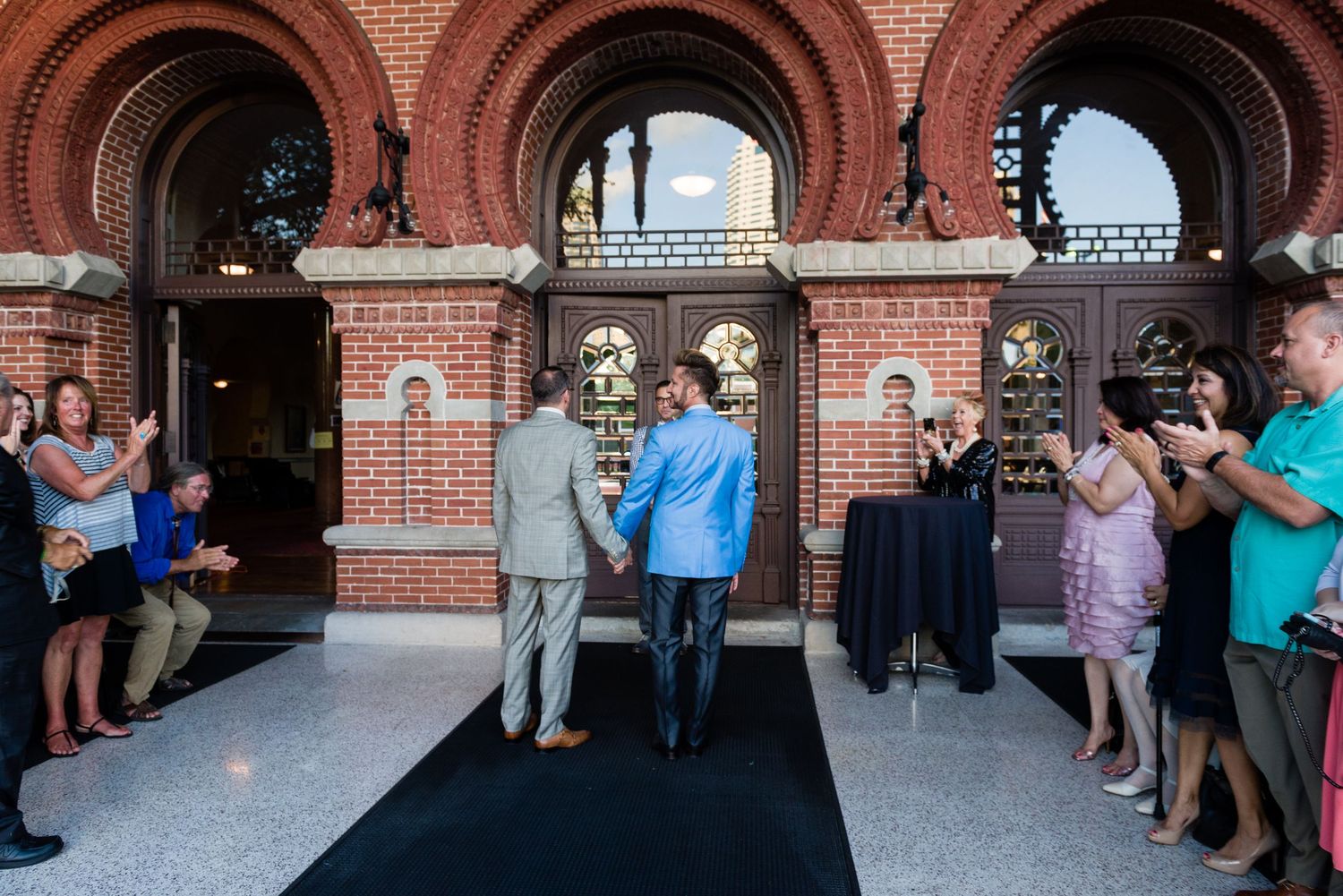 Two men shake hands on a black carpet outside a grand red brick building during an outdoor ceremony.