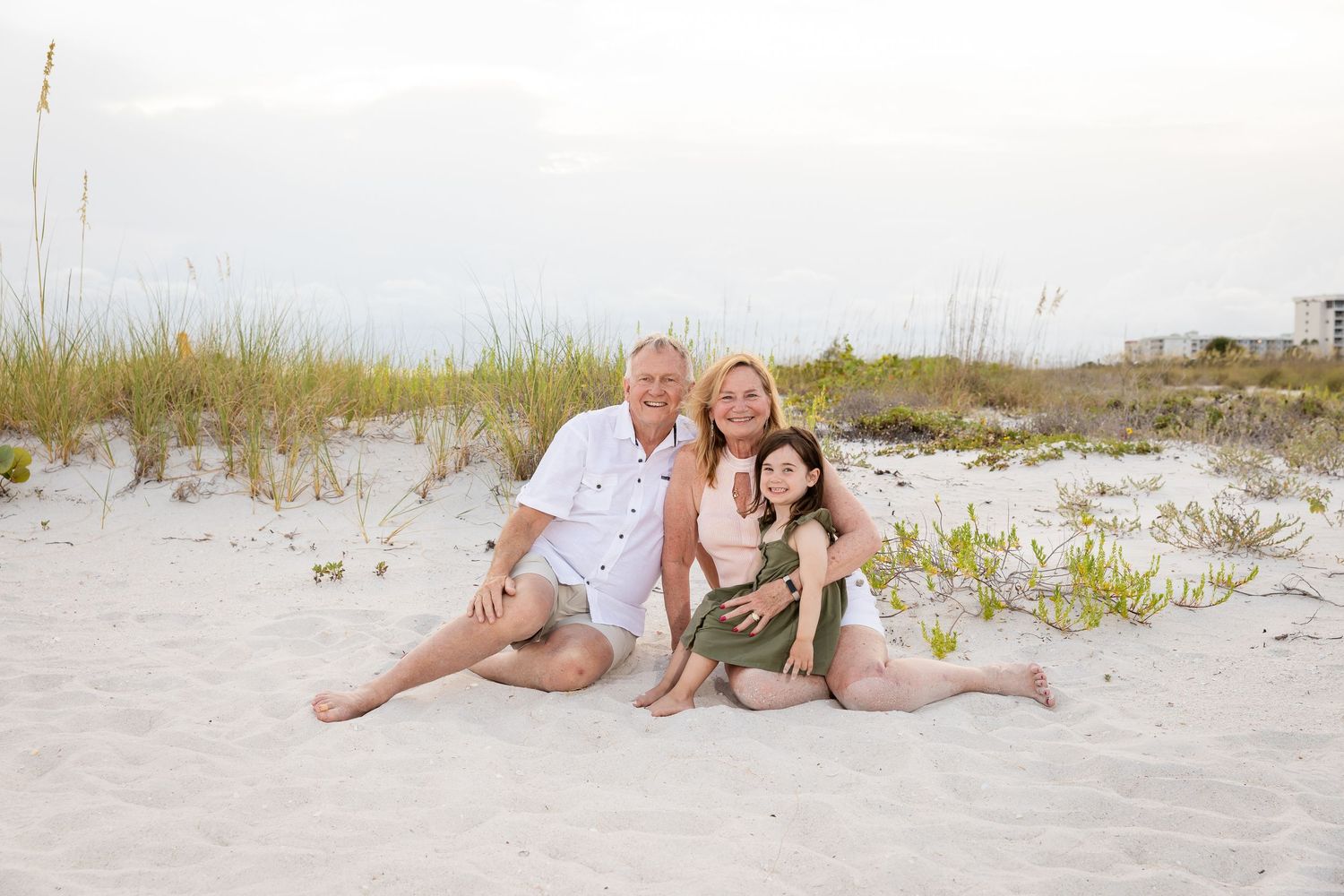 Family of three smiling together on a white sandy beach with sea oats and dunes in the background.