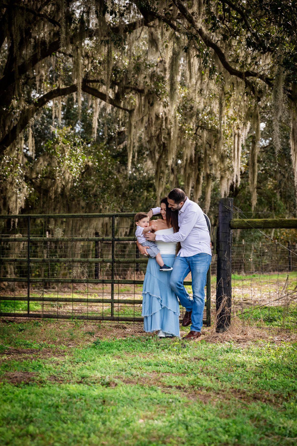 A loving couple kisses their baby near a black fence surrounded by Spanish moss-covered oak trees.