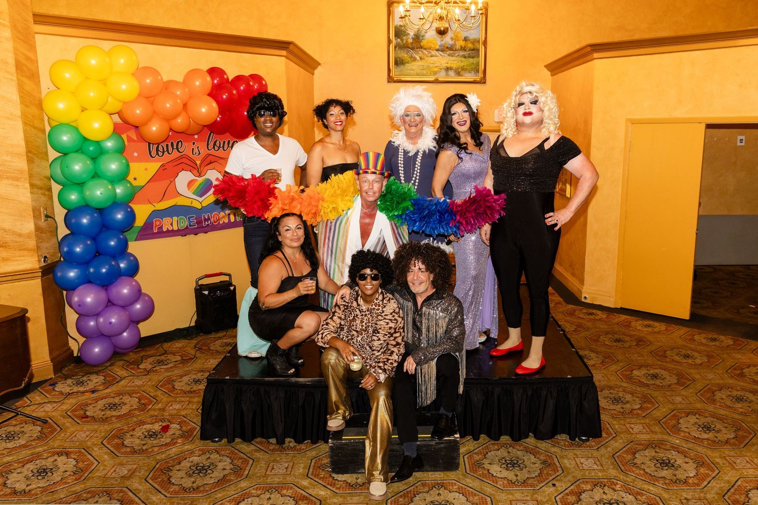 Diverse group of performers in colorful Pride costumes pose together in front of rainbow balloon arch backdrop.