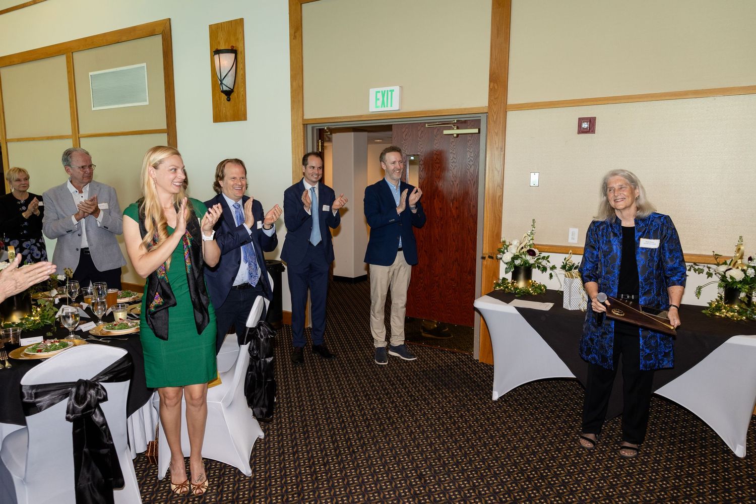 Crowd applauding a woman holding an award at a formal indoor banquet event with decorated tables.