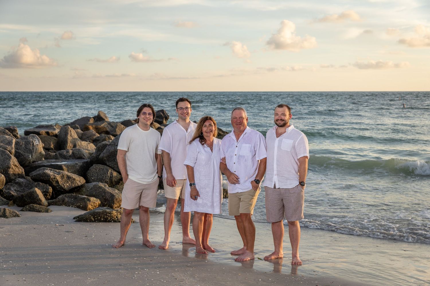 Family of five posing on a sandy beach at sunset near rocky jetty, wearing coordinated white and tan outfits.