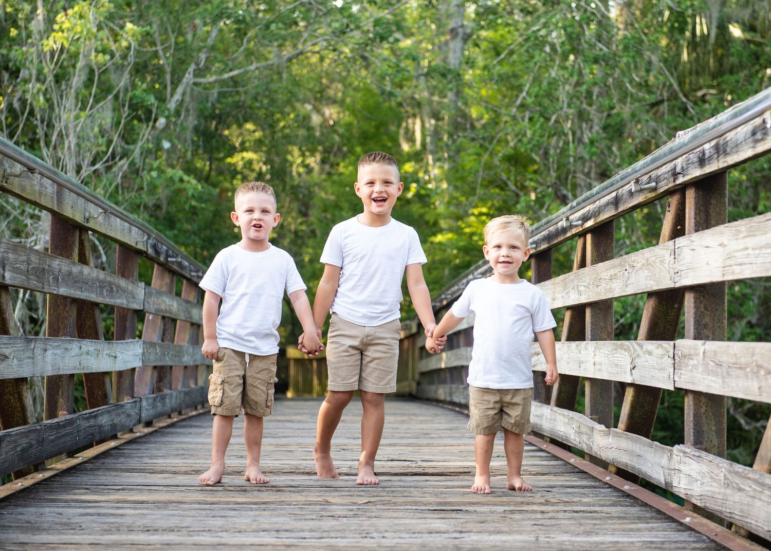 Three young boys holding hands and laughing on a wooden bridge surrounded by lush green trees.