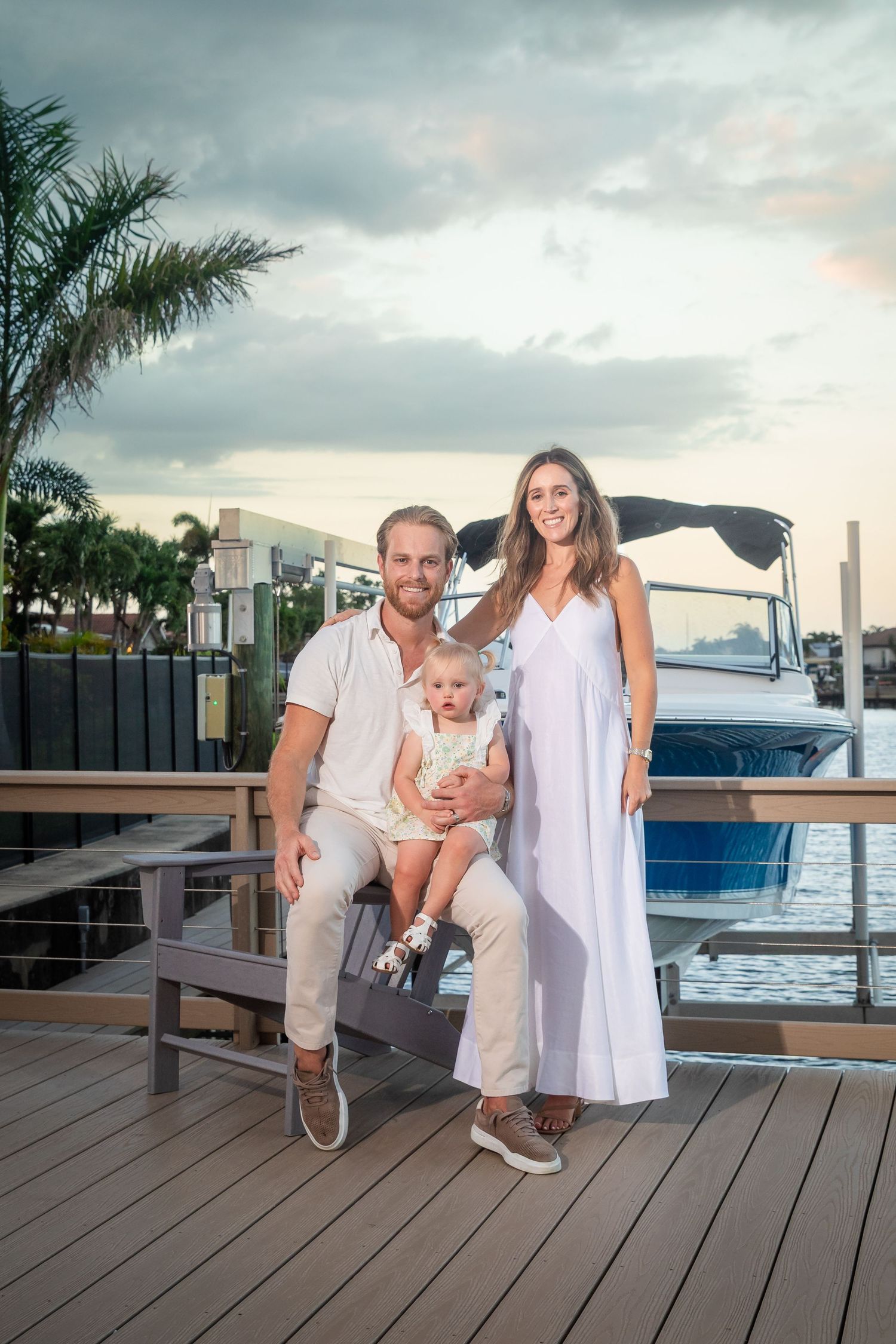 A smiling family of three poses on a waterfront dock at sunset, with a boat and palm trees in the background.