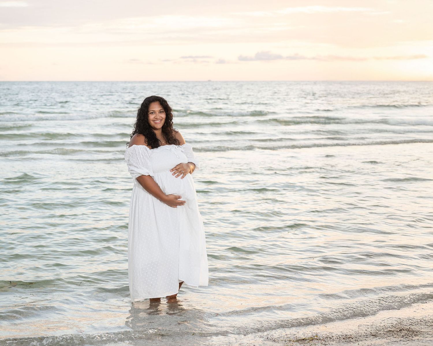 Pregnant woman in white off-shoulder dress standing in ocean waves at sunset beach, cradling baby bump.