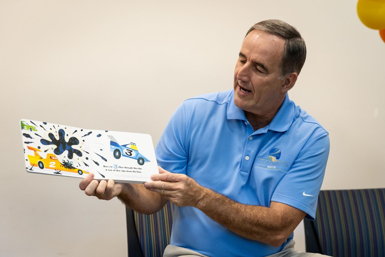 Man in blue polo shirt reads colorful children's book, smiling while holding it open to display illustrated pages.