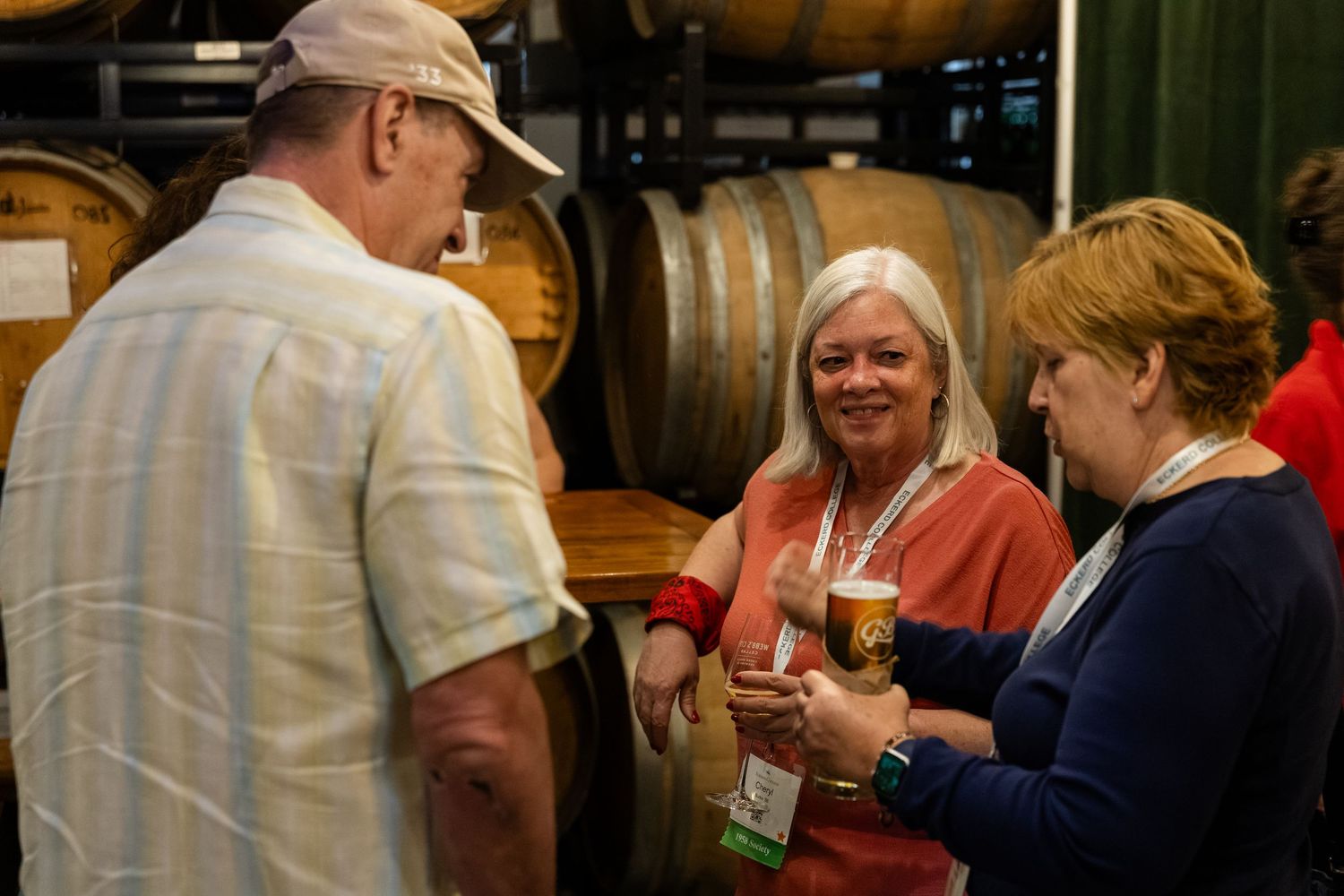 Three people socializing at a brewery tasting event with wooden barrels in the background.