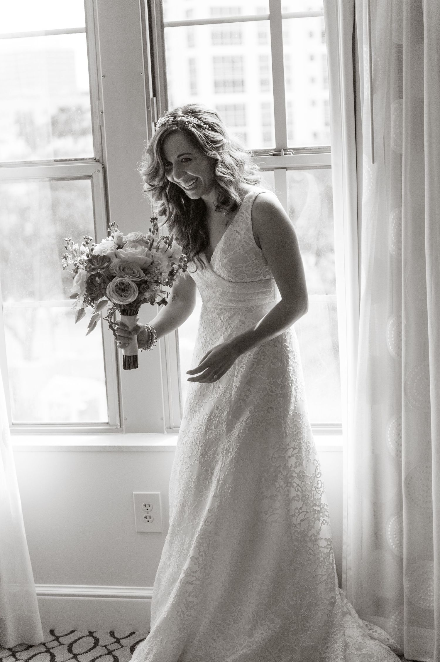 Black and white portrait of a radiant bride in a lace wedding gown holding a bouquet by a bright window.