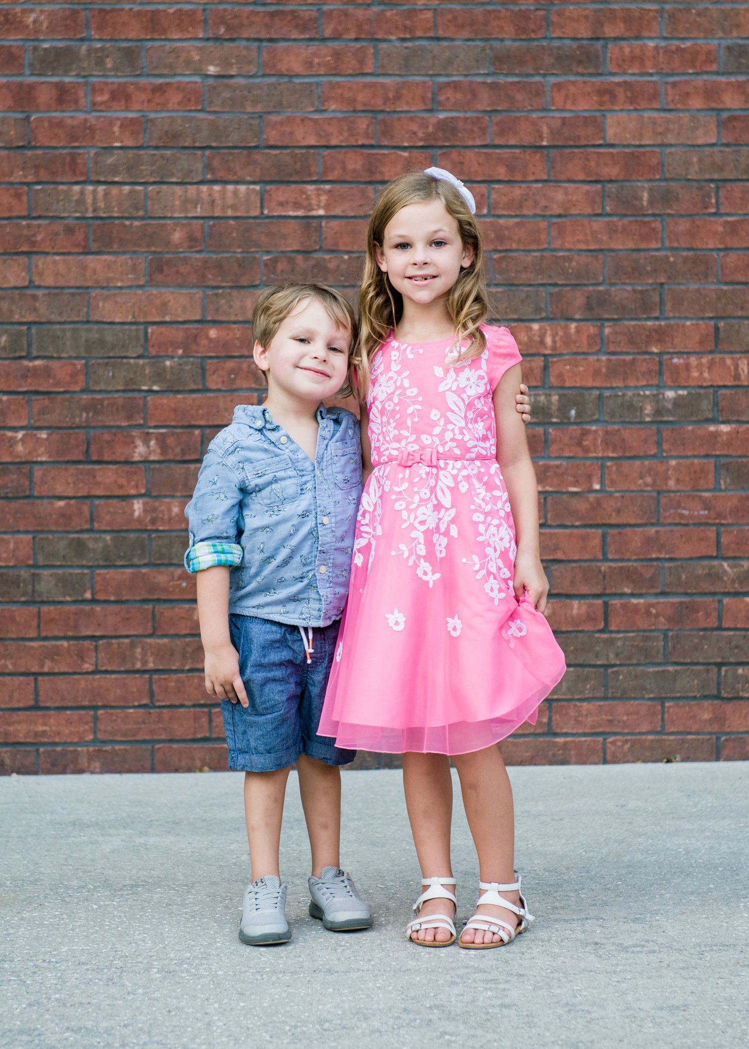 Two young children posing together in front of a brick wall, boy in blue shirt and girl in pink floral dress.