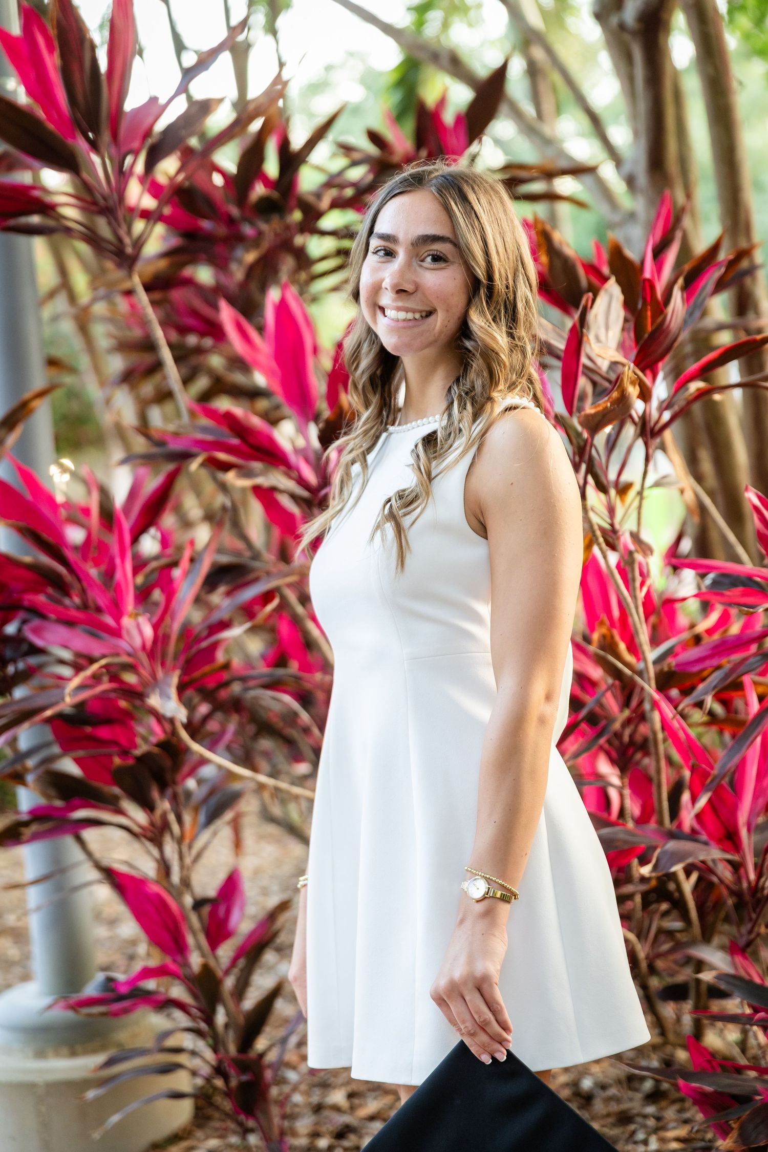 Young woman in white sleeveless dress smiling among vibrant red tropical plants outdoors.